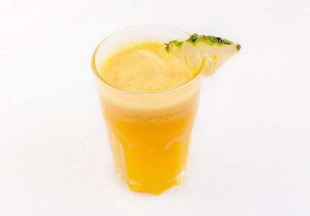 Copo de suco de abacaxi fresco com espuma natural e uma rodela da fruta na borda, posicionado sobre uma mesa de madeira num ambiente iluminado.