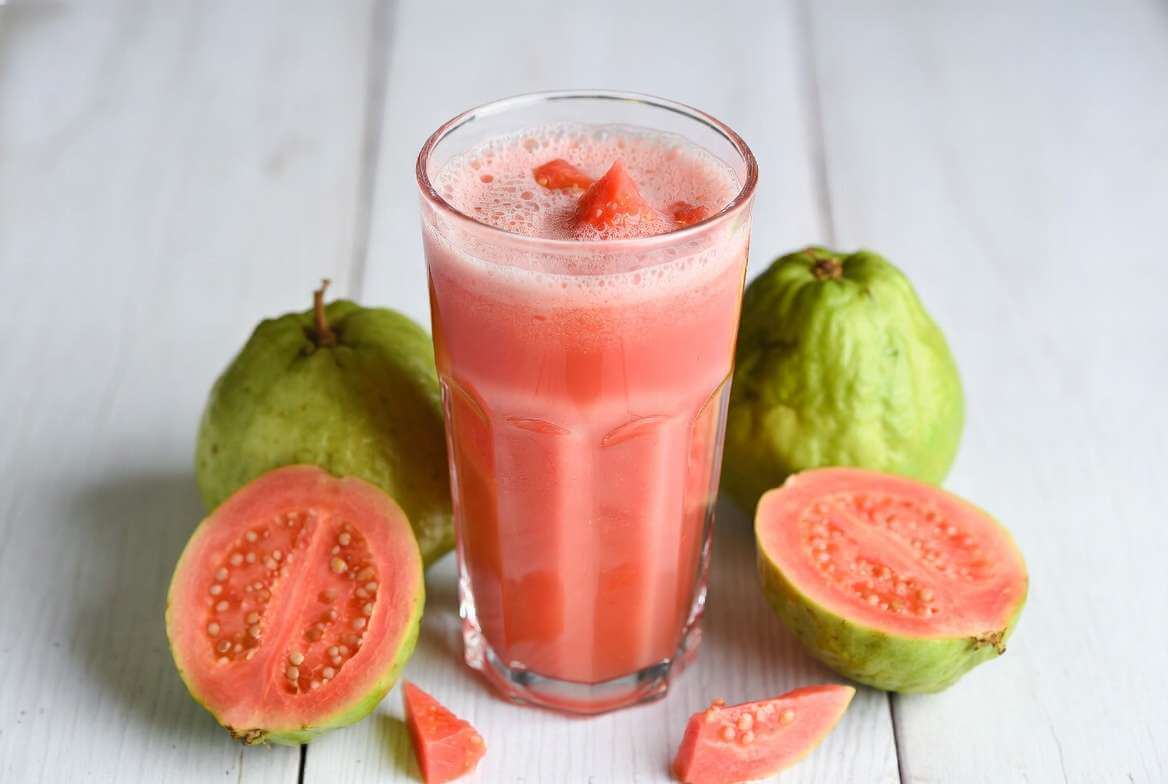 Copo de vidro transparente com suco de goiaba vermelha ao lado de uma goiaba fresca cortada ao meio, exibindo a polpa e as sementes.
