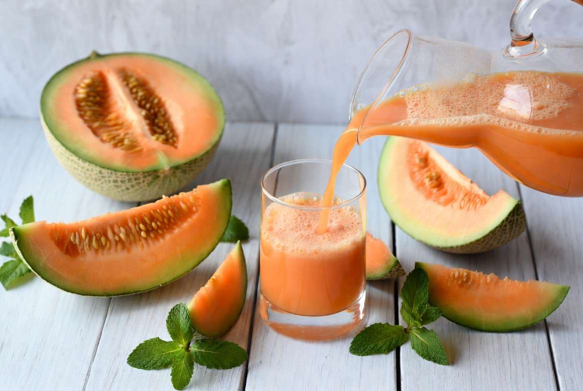 Um copo com suco de melão amarelo posicionado ao lado de um melão cortado ao meio, mostrando as sementes sobre a mesa.