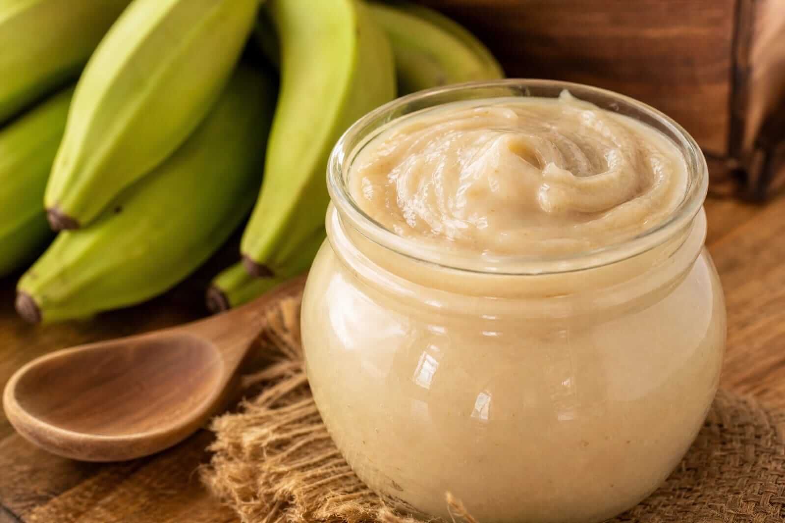 Pote de vidro com biomassa de banana verde cremosa posicionado ao lado de bananas verdes inteiras com casca.