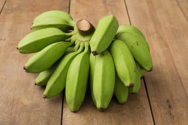 Banana-Figo Verde: Benefícios, Receitas e Como Fazer 3 Detalhe aproximado de um cacho de banana-figo verde destacando a casca grossa e as arestas quadradas da fruta.