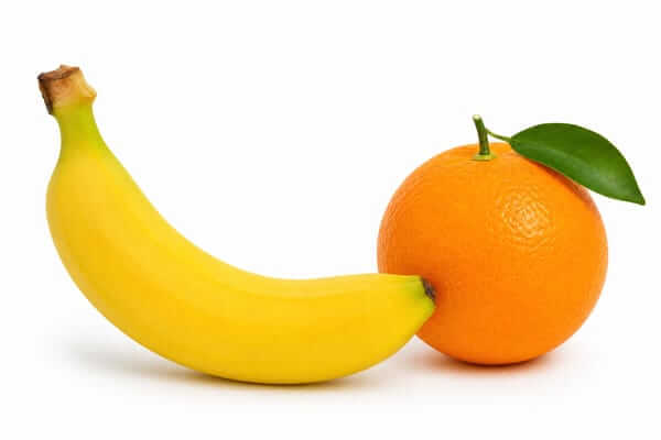 Imagem em plano aproximado com fundo branco minimalista. Em grande plano, estão uma banana e laranja inteiras, deitadas lado a lado. A banana tem a casca amarela e madura, e a laranja exibe uma casca cor de laranja texturada e brilhante.