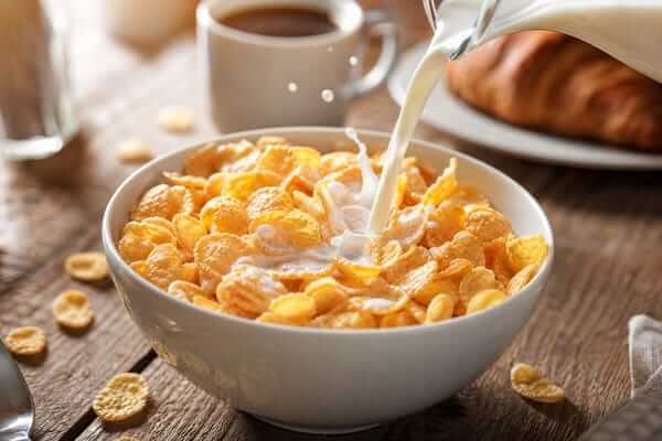 Cereal matinal: Energia e praticidade na sua manhã 1 Uma vista superior de uma taça de cerâmica branca cheia de cereal matinal crocante em flocos de trigo integral, no momento exato em que um fluxo contínuo de leite fresco é derramado por cima, criando pequenos salpicos.