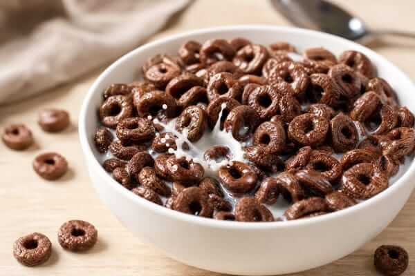 Cereal de chocolate: Mais sabor e humor na sua rotina 2 Uma taça de iogurte grego natural coberta com sucrilhos de chocolate crocantes, sementes de chia e morangos frescos cortados ao meio, disposta numa mesa de madeira iluminada pela luz do sol matinal