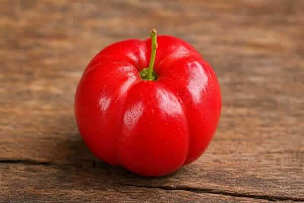Acerola: Benefícios da Fruta que Fortalece a Imunidade 2 "Uma acerola vermelha e madura posicionada no centro de uma tábua plana de madeira, num cenário limpo e focado no ingrediente."