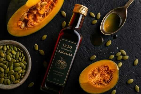 Um frasco de vidro escuro ao lado de uma colher de sopa com o líquido verde-escuro do óleo de semente de abóbora sobre uma mesa.
